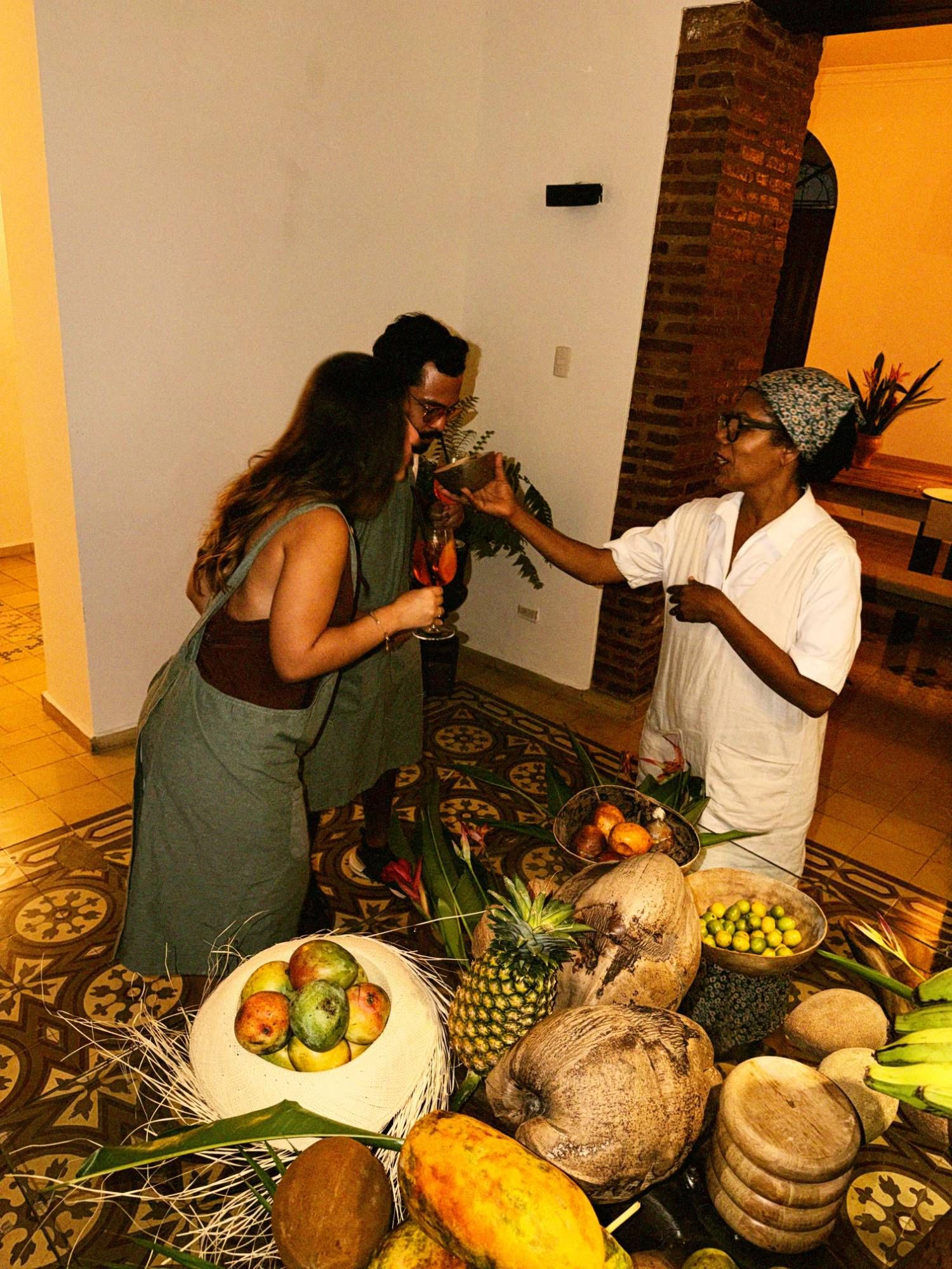 La cocinera de la casa, con pañuelo y bata blanca, da a oler un coco a dos invitados con delantal, junto a una mesa de frutas tropicales sobre el piso de mosaicos