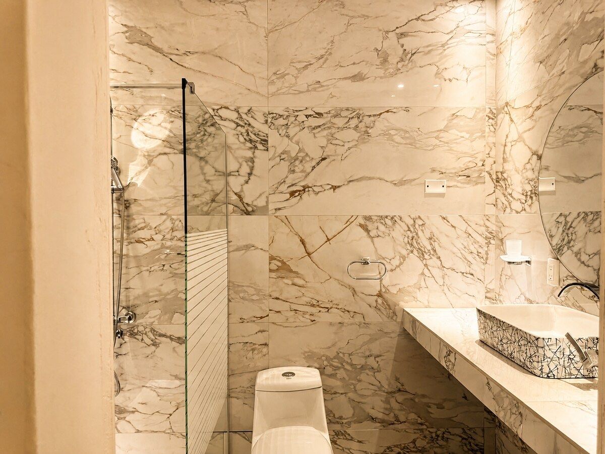 El baño privado, de mármol blanco, con ducha de vidrio