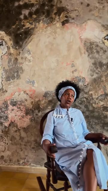 Una mujer con turbante, sentada en una mecedora frente a una pared antigua de la casa, habla a cámara