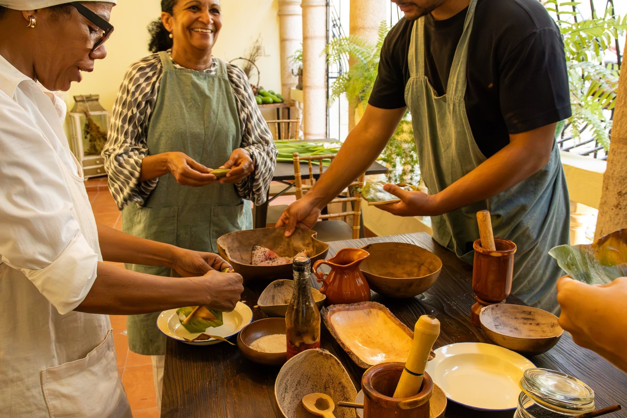 Maribel cocina con dos invitados en la mesa del patio, entre vasijas de madera, pilones y hojas de plátano